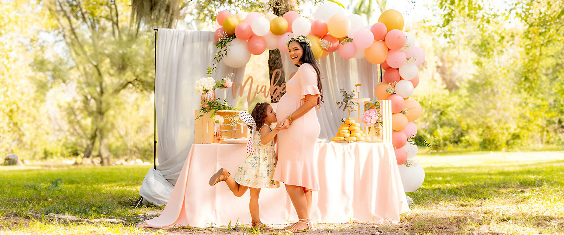 A woman at a baby shower with decorations behind, and child kissing her bump