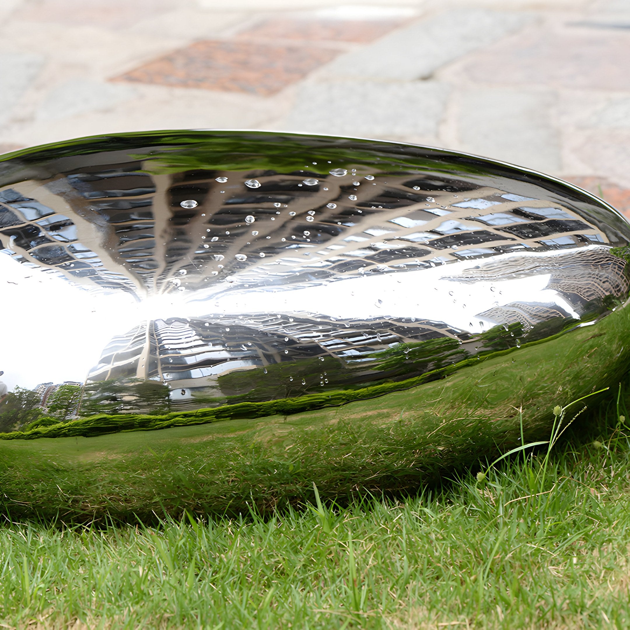 Close-up of polished stainless steel pebble sculpture on grass, showing reflective surface with water droplets.